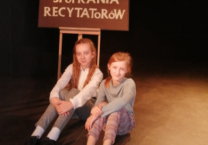 Dwie dziewczynki siedzą przy plakacie "Jesienne Spotkania Recytatorów"