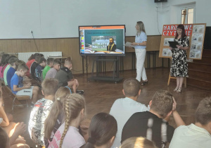 Nauczycielki języka polskiego przemawiają z prezentacją przed dziećmi w auli.
