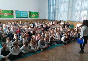 Dzieci siedzą w auli i uczestniczą w strojach galowych w lekcji otwartej.